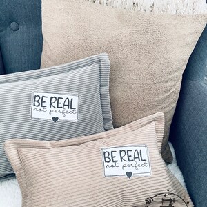 以下が含まれることがあります： 「BE REAL not perfect」と刺繍されたベージュのクッションが2つ。クッションは、白と黒の毛布がかけられた濃い青色のソファの上に置かれています。