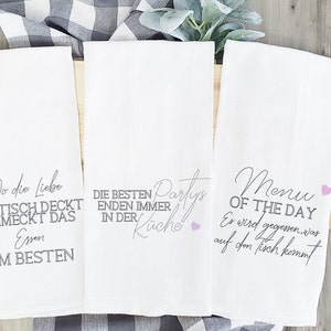 May include: Three white kitchen towels with pink hearts and gray text. The first towel says "Wo die Liebe den Tisch deckt schmeckt das Essen am besten". The second towel says "Die besten Partys enden immer in der Küche". The third towel says "Menu of the Day Er wird gegessen, was auf den Tisch kommt".