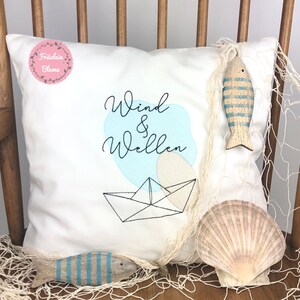 Op de afbeelding: Wit kussen met een blauw en beige geborduurd ontwerp van een papieren boot en de tekst "Wind & Wellen". Het kussen ligt op een houten stoel met een visnet en twee houten vissen met blauwe strepen.