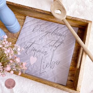 May include: A gray quilted pot holder with the text "Die geheime Zutat ist immer Liebe" embroidered in black. The pot holder is sitting on a wooden tray with a wooden spoon.