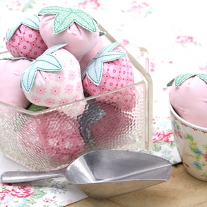 Può includere: Una ciotola di vetro piena di fragole di stoffa rosa e bianca. Le fragole hanno foglie verdi e sono disposte in un motivo decorativo. Un mestolo d'argento è accanto alla ciotola. Una tazza da tè bianca con un motivo floreale contiene una fragola di stoffa rosa.