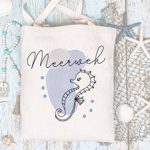 Op de afbeelding: Een crèmekleurige draagtas met een illustratie van een zeepaardje en het woord "Meerweh" in zwart schrift. Het zeepaardje is zwart omlijnd, geplaatst tegen een blauw hartvormig ontwerp. De tas heeft beige handvatten en is van canvas.