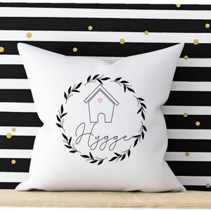 Könnte beinhalten: Ein weißes Kissen mit einem schwarz-weißen Design eines Hauses mit einem Herzen darin, umgeben von einem Kranz aus Blättern und dem Wort "Hygge" in Kursivschrift.