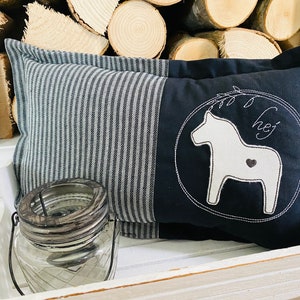 May include: A black and white striped cushion with a black cushion featuring a white embroidered Dala horse with a heart and the word "hej" in a circle. The cushions are sitting in a white wooden tray with a clear glass jar.