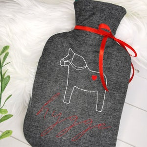 Pode incluir: Bolsa de água quente de tecido cinza com uma fita vermelha e um cavalo Dala bordado em branco com um coração vermelho. O texto "Hygge" está bordado em vermelho.
