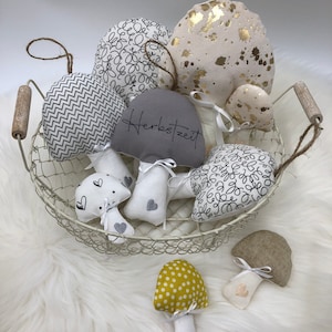 Pode incluir: Uma coleção de enfeites decorativos em forma de cogumelo numa cesta de arame. Os cogumelos apresentam vários padrões, incluindo bolinhas, espirais e chevron, com cores como branco, cinza e dourado. Alguns têm a palavra "Herbstzeit" bordada.