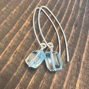 Puede incluir: Un par de pendientes de plata con gotas de piedras preciosas azul claro. Los pendientes están hechos con un diseño simple de alambre y presentan una piedra preciosa de forma rectangular.
