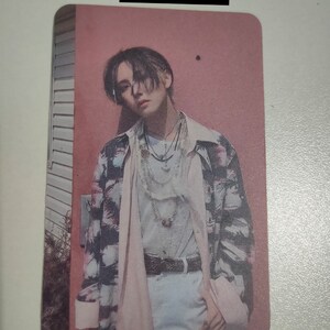 May include: A rectangular trading card with a matte coat finish featuring a portrait of a person with dark hair wearing a patterned button-down shirt over a light-colored top, layered necklaces, and a belt, set against a pink wall background.