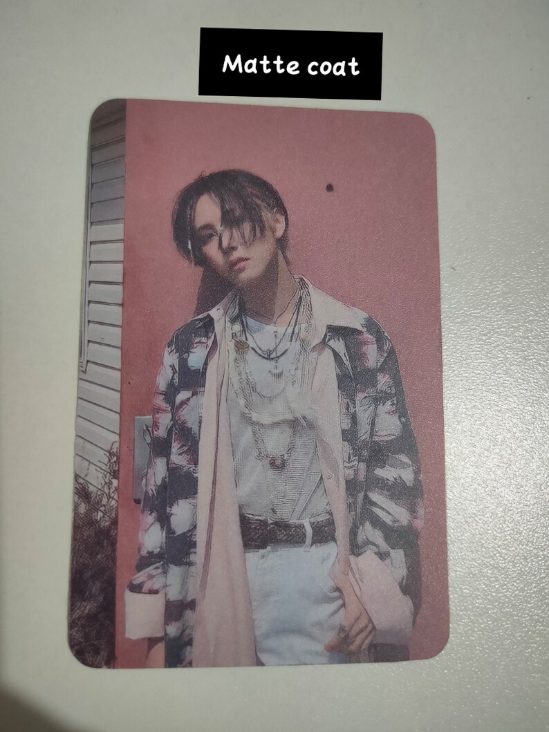 May include: A rectangular trading card with a matte coat finish featuring a portrait of a person with dark hair wearing a patterned button-down shirt over a light-colored top, layered necklaces, and a belt, set against a pink wall background.