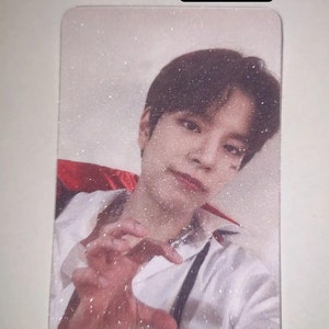 May include: A rectangular photo card featuring a person with dark hair wearing a white shirt with a red collar accent. The image has a glittered finish and displays the text "Glittered" in a black box at the top. The person is making a claw gesture.