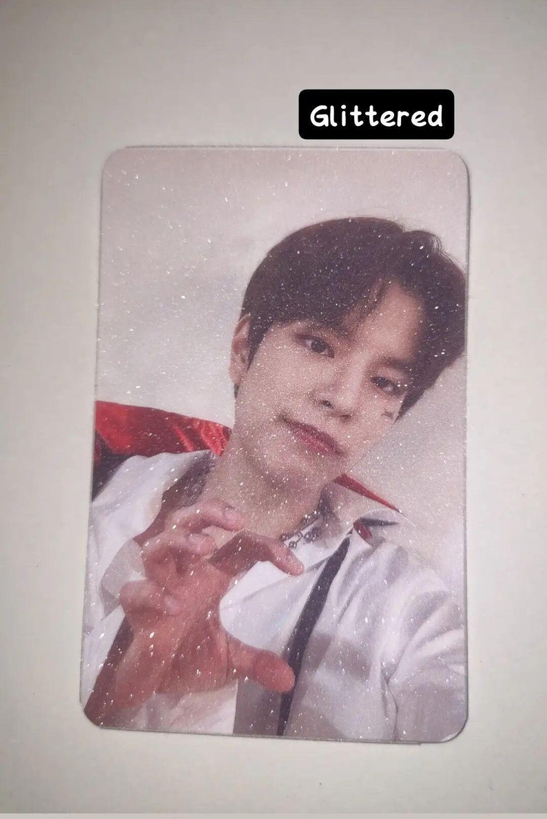 May include: A rectangular photo card featuring a person with dark hair wearing a white shirt with a red collar accent. The image has a glittered finish and displays the text "Glittered" in a black box at the top. The person is making a claw gesture.