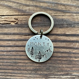 Könnte beinhalten: Ein silberner Schlüsselanhänger mit einem runden Anhänger, auf dem "MONTY." steht. Der Anhänger zeigt ein Design mit drei Bäumen und Bergen.