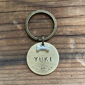 Könnte beinhalten: Ein Messing-Hundeanhänger mit einem Knochen-Anhänger und dem eingravierten Namen "YUKI". Der Anhänger hat ein Herz und Pfotenabdrücke um den Namen herum.