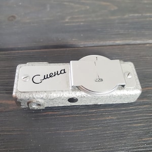 May include: A vintage silver-toned camera with the Cyrillic word "Смена" on the front. The camera has a textured body and a circular dial with a needle and the number 1. A small lens is visible on the side.