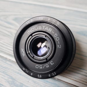 May include: A black Industar-50-2 camera lens with a series of numbers and markings around the edge. The lens has a glass element in the center and the text "INDUSTAR-50-2" is visible.