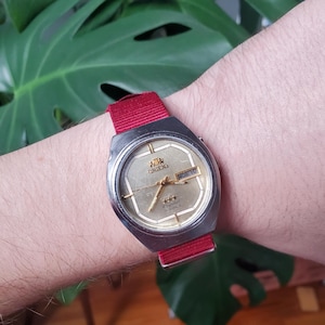 Może przedstawiać: Zegarek Orient Crystal w stylu vintage ze srebrną kopertą i złotą tarczą. Zegarek posiada czerwony materiałowy pasek i wyświetla dzień tygodnia. Zegarek jest noszony na nadgarstku.