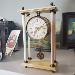 May include: A vintage ivory and gold-toned mantel clock. The clock features a round face with Roman numerals, gold hands, and a decorative pendulum. The clock is supported by four ivory columns and a rectangular base.
