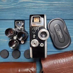 Puede incluir: Una cámara vintage en blanco y negro con la palabra "Эkран" en escritura. La cámara va acompañada de una lente, un visor, una tapa de lente y una funda de cuero marrón. Los objetos están dispuestos sobre una superficie de madera azul.