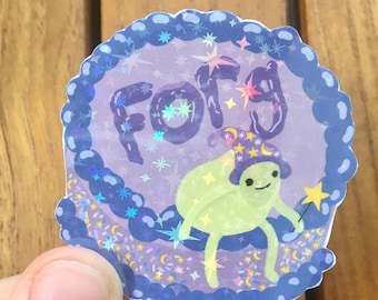 May include: A round sticker with a purple border and a light purple background. The sticker features the word "FORG" in purple, a green cartoon character wearing a wizard hat, and a yellow star wand. The sticker has a holographic effect.
