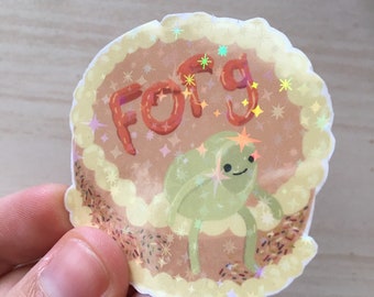 May include: A brown and yellow sticker with a green frog and the word "FORG" in red frosting. The sticker has a glittery, iridescent finish.