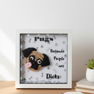 May include: A white framed artwork with a crocheted pug face. The background has paw prints and the text "Pugs because people are Dicks". The pug has black ears and a pink tongue.
