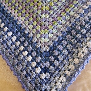 May include: A square crochet blanket featuring a granny square pattern in variegated shades of blue, purple, green, and white. The textile has a textured surface composed of repeating clusters of yarn stitches with a scalloped border edge.