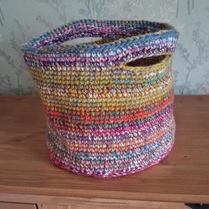 May include: A round, hand-crocheted basket featuring a vibrant, multi-coloured yarn pattern with horizontal stripes of pink, orange, yellow, blue, and purple. The basket has a thick, textured stitch and two integrated side handles.