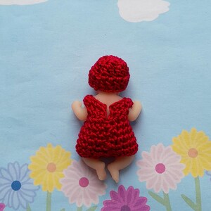 May include: A handmade doll with a red crocheted dress and matching cap. The doll is lying on a blue background with white clouds and floral patterns. The doll's skin is a light beige color.
