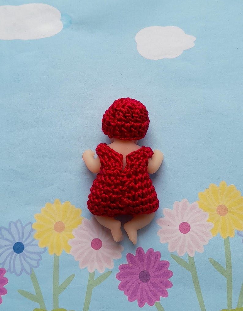 May include: A handmade doll with a red crocheted dress and matching cap. The doll is lying on a blue background with white clouds and floral patterns. The doll's skin is a light beige color.