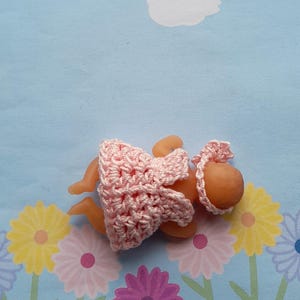 May include: A small doll dressed in a handmade pink crochet dress and bonnet. The doll is lying on a blue floral patterned background. The crochet work is delicate and detailed.