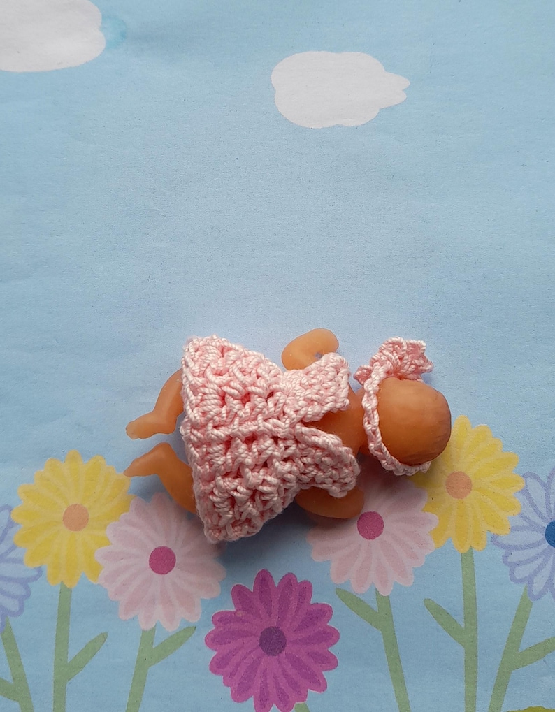 May include: A small doll dressed in a handmade pink crochet dress and bonnet. The doll is lying on a blue floral patterned background. The crochet work is delicate and detailed.
