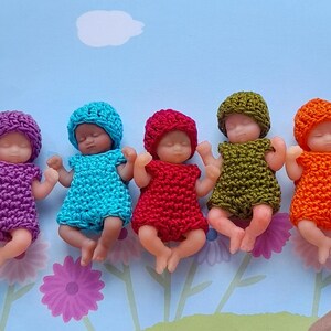 May include: Five miniature baby dolls, each wearing a crocheted outfit and matching hat. The outfits are in shades of purple, blue, red, olive green, and orange. The dolls are lying on a light blue surface with floral and bird designs.