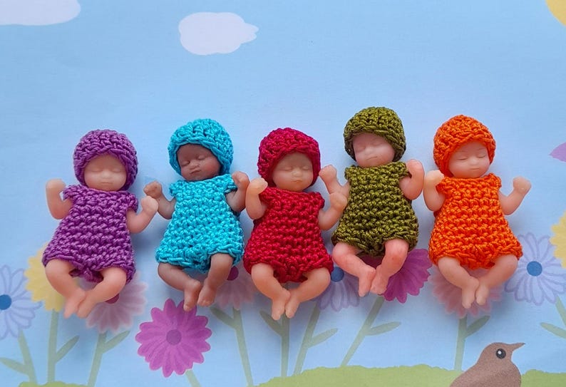 May include: Five miniature baby dolls, each wearing a crocheted outfit and matching hat. The outfits are in shades of purple, blue, red, olive green, and orange. The dolls are lying on a light blue surface with floral and bird designs.