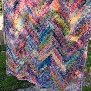 May include: A rectangular crochet blanket featuring a vibrant chevron pattern. The textile is composed of multicoloured yarn in shades of pink, blue, green, yellow, and purple, worked in a dense, textured stitch with a finished border.