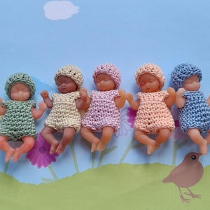 以下が含まれることがあります： 緑、黄色、ピンク、ピーチ、青のパステルカラーの、かぎ針編みの衣装と帽子を着た5体の小さな赤ちゃん人形。人形は、花と鳥のデザインが施された青い背景の上に横たわっています。