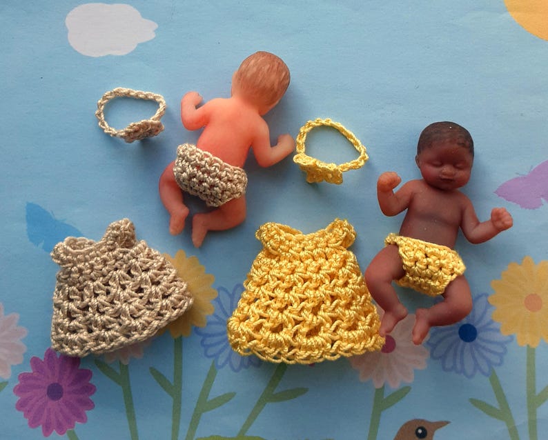 May include: Miniature baby dolls with crocheted outfits and headbands. The dolls have different skin tones and wear beige and yellow crocheted dresses and nappies. The background is a light blue surface with floral patterns.