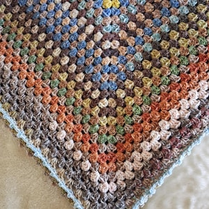 May include: A square crochet blanket featuring a multi-coloured granny square pattern. The design incorporates shades of brown, blue, yellow, green, and orange yarn, finished with a light blue scalloped border along the outer edge.