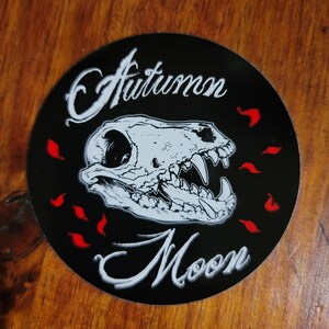 May include: A black and white sticker with the text "Autumn Moon" in a stylized font. The sticker features a white animal skull with red leaf shapes around it.