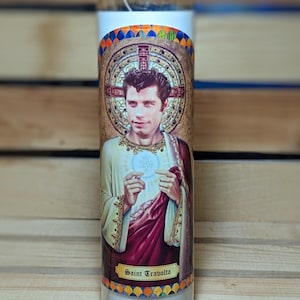 May include: A white candle with a colorful image of John Travolta as a saint. The image is surrounded by a gold frame and the text "Saint Travolta" is printed below the image.