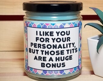 May include: A glass jar candle with a black lid and light blue wax. The label reads "I LIKE YOU FOR YOUR PERSONALITY, BUT THOSE TITS ARE A HUGE BONUS" with a colorful geometric border. A small potted succulent is to the right.