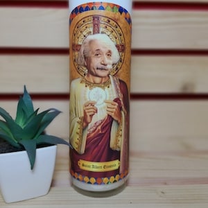 May include: A white candle with a colorful image of Albert Einstein on the front. The image depicts Einstein in a robe with a halo and a cross behind him. The text "Saint Albert Einstein" is printed below the image.