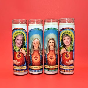 May include: Four white pillar candles with colorful images of women and text. The candles are labeled "Saint Rose Nylund", "Saint Sophia Petrillo", "Saint Dorothy Zbornak", and "Saint Blanche Devereaux".