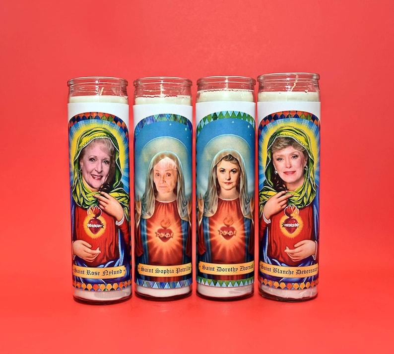 May include: Four white pillar candles with colorful images of women and text. The candles are labeled "Saint Rose Nylund", "Saint Sophia Petrillo", "Saint Dorothy Zbornak", and "Saint Blanche Devereaux".