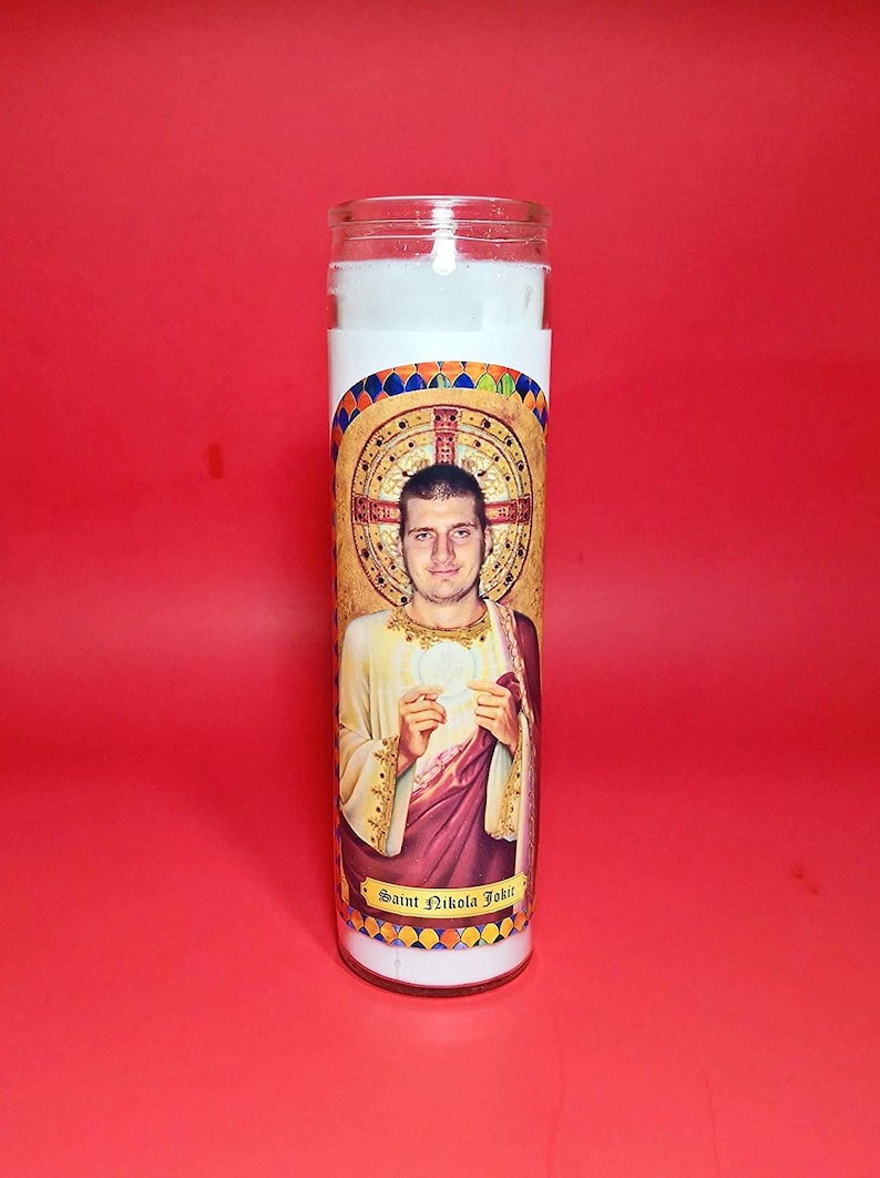 May include: A white candle with a colorful image of a man with a halo and the text "Saint Nikola Jokic".