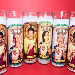 May include: Six white pillar candles with colorful images of the six main characters from the TV show Friends. Each candle has a gold crown and the name of the character below the image. The names are "Saint Joey", "Saint Phoebe", "Saint Ross", "Saint Monica", "Saint Rachel", and "Saint Chandler".