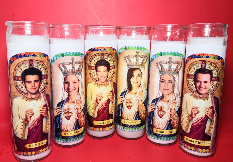 May include: Six white pillar candles with colorful images of the six main characters from the TV show Friends. Each candle has a gold crown and the name of the character below the image. The names are "Saint Joey", "Saint Phoebe", "Saint Ross", "Saint Monica", "Saint Rachel", and "Saint Chandler".