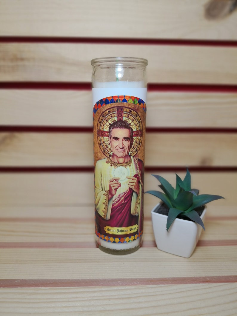May include: A white candle with a colorful image of a man with a halo and the text "Saint Johnny Rose".