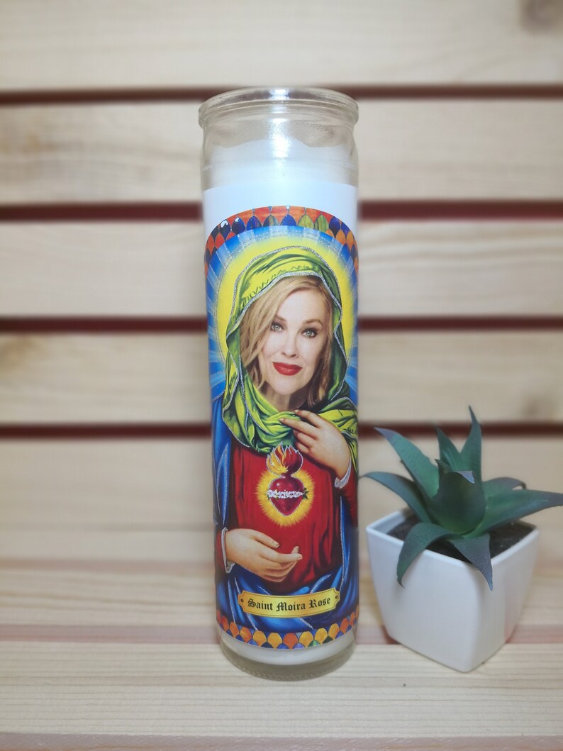 May include: A white candle with a colorful image of a woman with a heart on her chest. The text "Saint Moira Rose" is printed on the bottom of the candle.