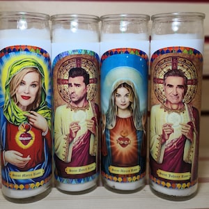 May include: Four white pillar candles with colorful images of people and text. The candles are labeled "Saint Moira Rose", "Saint David Rose", "Saint Alexis Rose", and "Saint Johnny Rose".