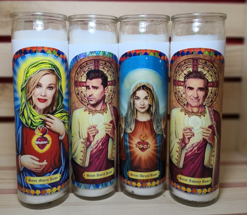 May include: Four white pillar candles with colorful images of people and text. The candles are labeled "Saint Moira Rose", "Saint David Rose", "Saint Alexis Rose", and "Saint Johnny Rose".